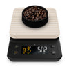 A digital kitchen scale with a white cover and black matte finish, featuring a digital display showing weight and timer, with a bowl of coffee beans on top.
