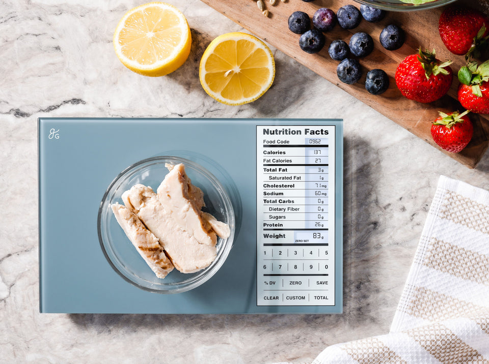 A small bowl of grilled chicken being weighed on the Nutrition Label Scale. Around it on the countertop are two lemon halves, strawberries, blueberries, and nuts.