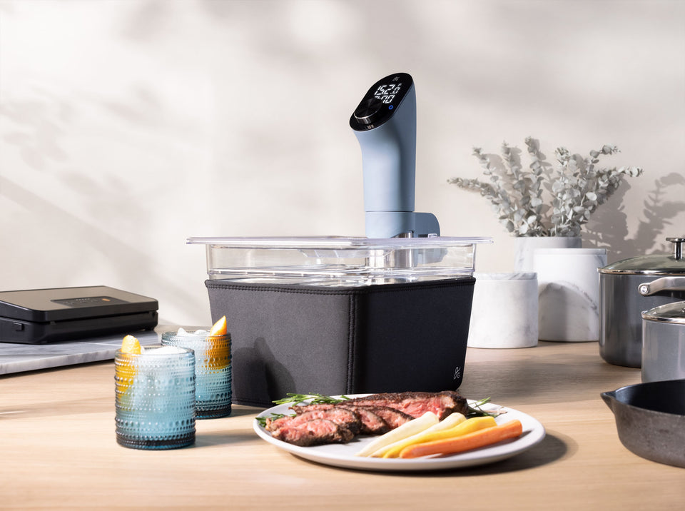 A sous vide clipped into the sous vide container with a black sleeve on it; they sit on a countertop next to a vacuum sealer, pair of drinks, and a plate of cooked steak and carrots.