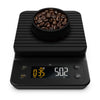 A digital kitchen scale with a black matte finish, featuring a digital display showing weight and timer, with a bowl of coffee beans on top.