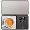 A digital pocket scale with a stainless steel weighing platform, open with an espresso cup on top, displaying a weight reading on its LCD screen.