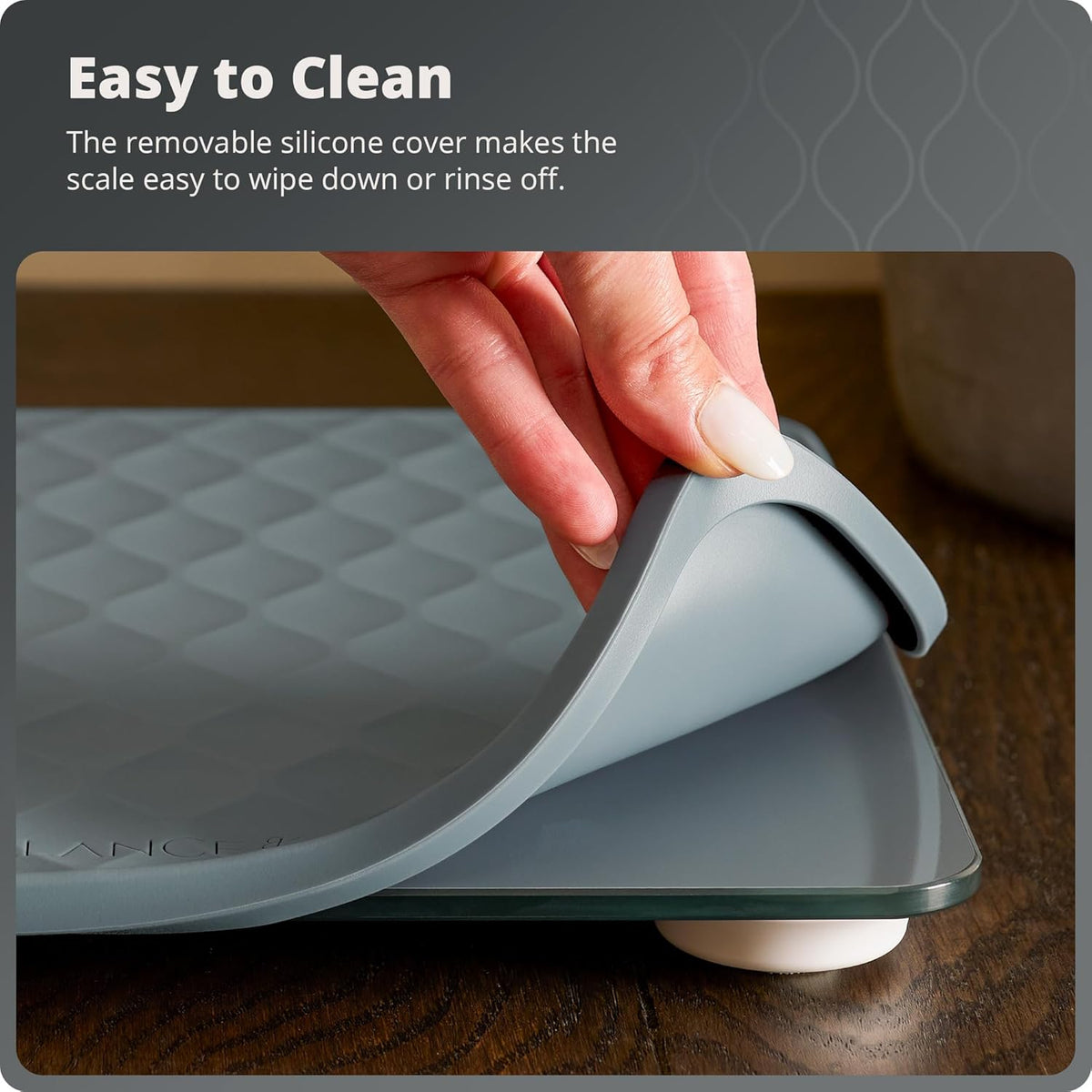 Gray scale with a removable silicone cover being held by a hand on a wooden surface. Text reads, "Easy to Clean: The removable silicone cover makes the scale easy to wipe down or rinse off."

