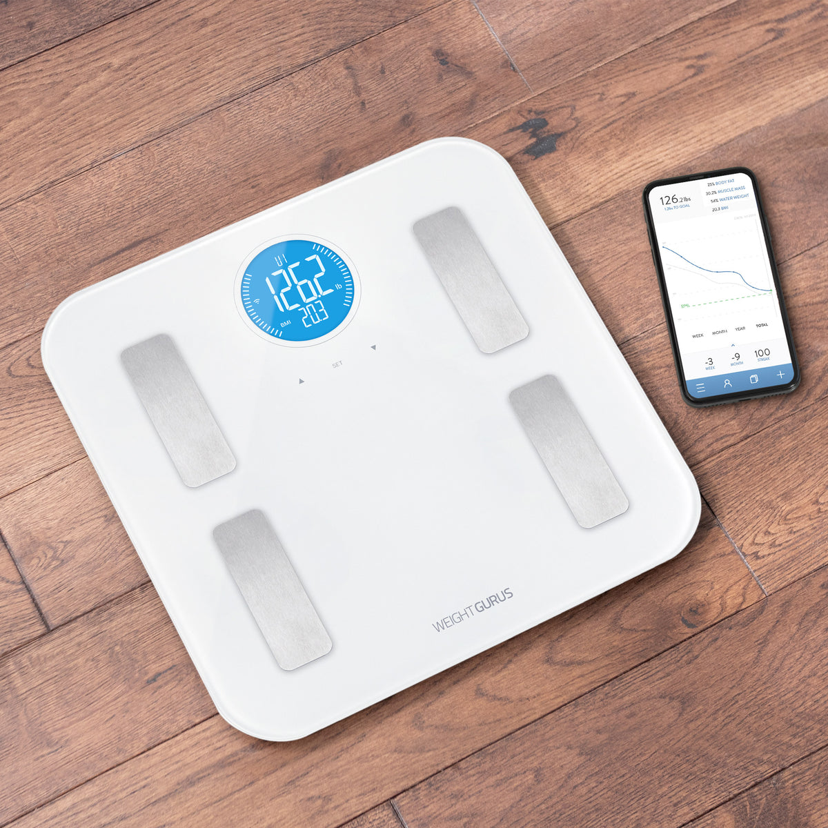 Greater Goods White WiFi Digital Body Composition Scale on the floor next to a phone displaying the Weight Gurus app. 