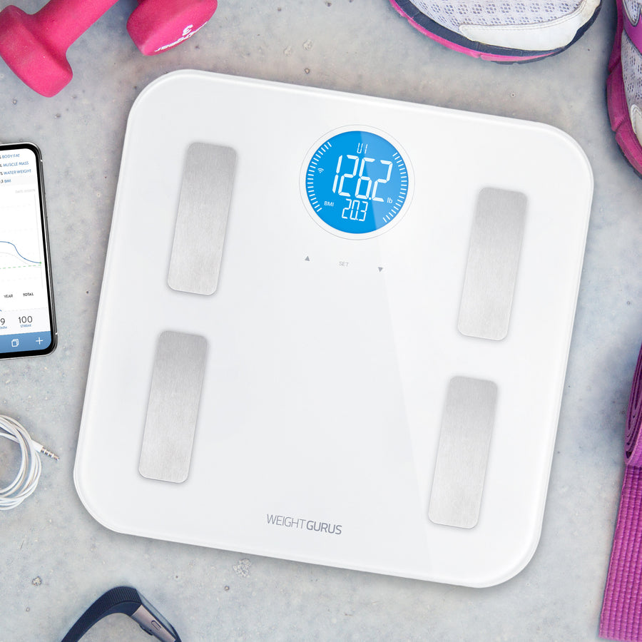 Greater Goods White WiFi Digital Body Composition Scale on the floor next to a phone, weights, and shoes.